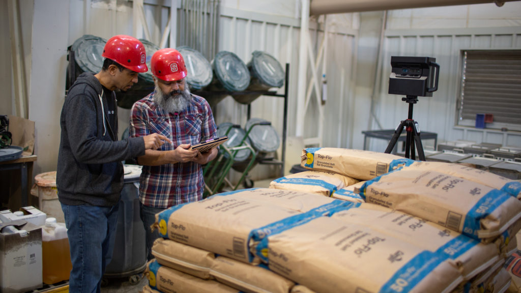 two men in red hard hats look at a tablet while standing in a feed mill